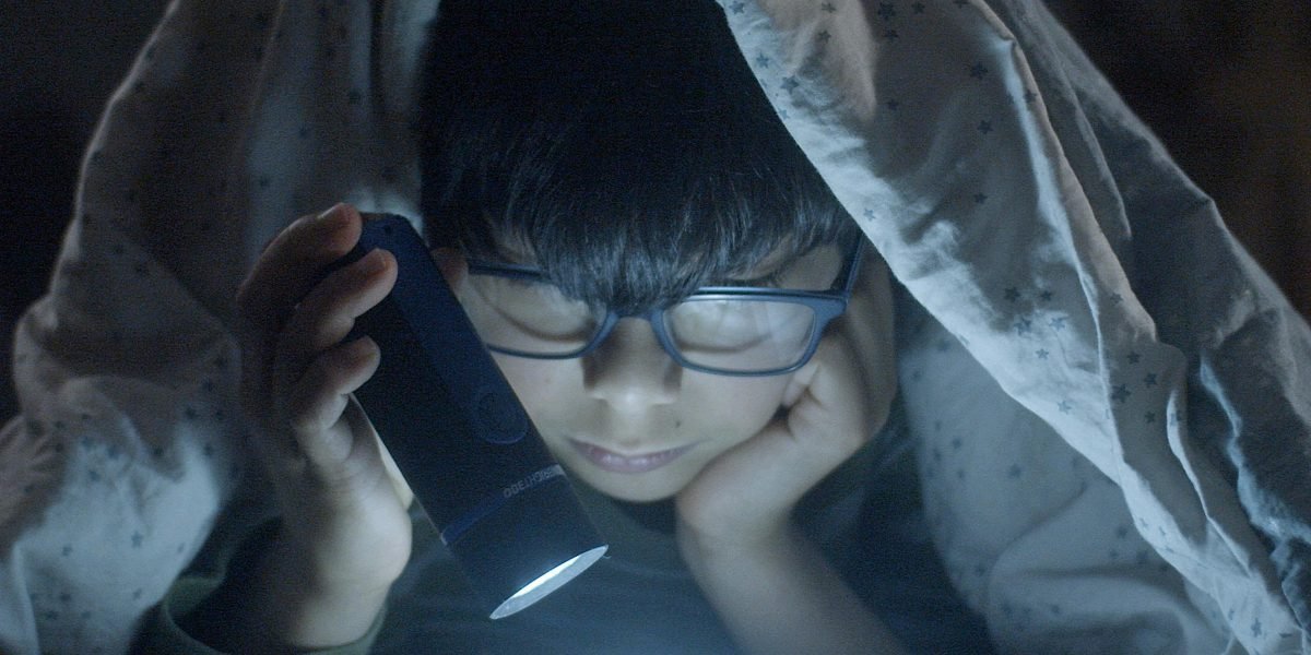 A child reading under blankets with a flashlight, evoking the nostalgic thrill of Goosebumps books.